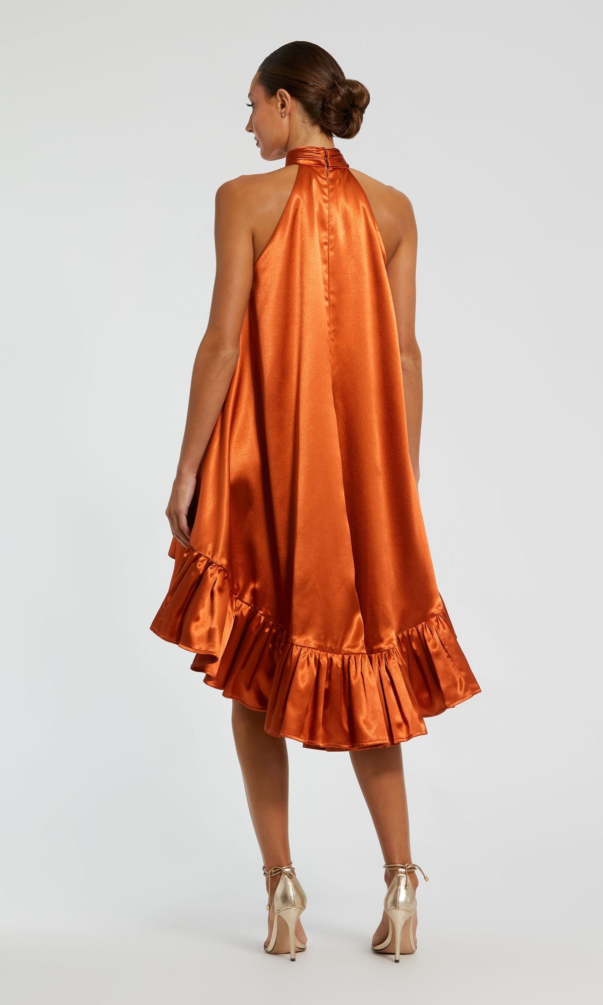 A woman faces away, wearing the Mac Duggal 20711 Satin Wedding Guest Dress in orange with a high-neck and ruffled high-low hemline, paired with metallic high-heeled shoes.